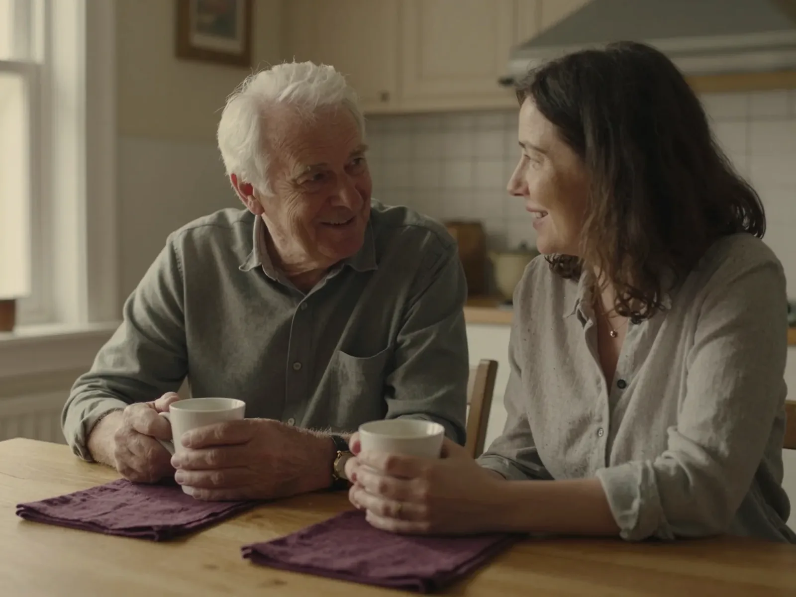 An older man and his adult daughter talking over coffee at a kitchen table.